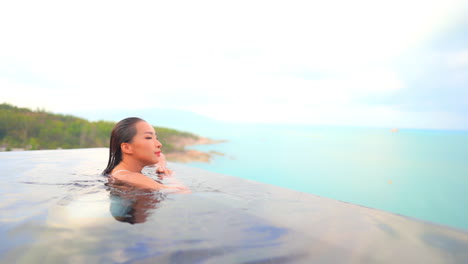 Asian-woman-swims-toward-edge-of-infinity-pool-overlooking-ocean-beauty-relaxing-poolside-in-fashion-swimsuit