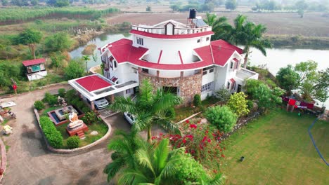 Zoom-out-over-palm-trees-and-birds-flying-at-the-relaxing-vacation-home-by-the-lake,-pool-and-parking-lot-in-Vadodara,-India