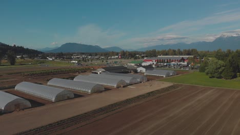 Local-harvest-farm-Chilliwack-BC-plastic-grow-tunnels-greenhouse-fields-row-vegetable-crops-in-the-beginning-of-winter-cars-trucks-on-highway-mountains-clouds-blue-sky-background-aerial-orbit-pan-up