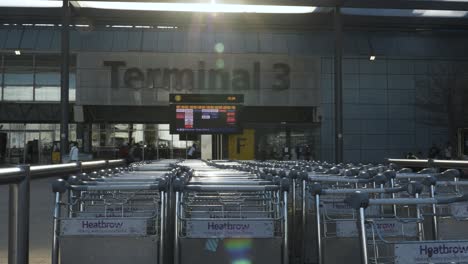 Fila-De-Carros-De-Equipaje-Fuera-Del-Edificio-De-La-Terminal-3-De-Heathrow-Con-Tablero-De-Salida-De-La-Aerolínea-Con-Bengalas-Solares