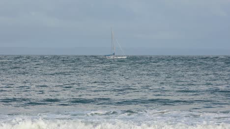 White-Sailboat-Sailing-In-Deep-Calm-Waters-with-some-waves-crashing-in-front-in-Carcavelos,-Cascais