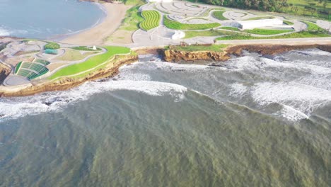 shot-taken-over-the-sea-slowly-zooming-into-an-expansive-structure-right-by-the-sea,-gentle-foamy-white-waves,-modern-architecture,-green-patch,-city-in-distance,-bright-day,-cliffside-visible,-rocky