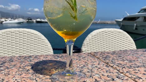 Fancy-cocktail-with-champagne-and-rosemary-by-the-sea-in-Puerto-Banus-with-yachts,-luxury-holiday-destination,-drink-on-a-tropical-vacation-with-sea-view-in-Marbella-Spain,-4K-shot