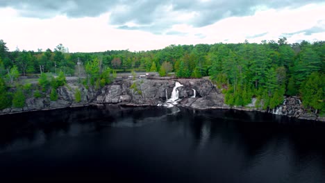 Gushing-waterfall-flows-down-rock-escarpment-into-dark-colored-lake-with-lush-green-forest-view-from-above-in-Northern-Ontario
