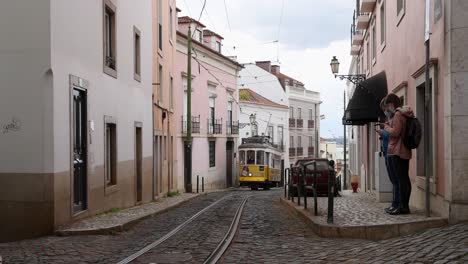 Zeitlupenansicht-Der-Berühmten-Straßenbahn-28-In-Lissabon,-Portugal,-Während-Sie-Eine-Schmale-Straße-Mit-Kopfsteinpflaster-Hinunterfährt,-Die-Von-Bunten-Gebäuden-Auf-Schienen-Und-Strom-Gesäumt-Ist