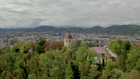 Aerial-push-towards-Glokenturm-tower-on-wooded-hilltop-with-Graz's-Schloßberg-Austria-mountain-skyline