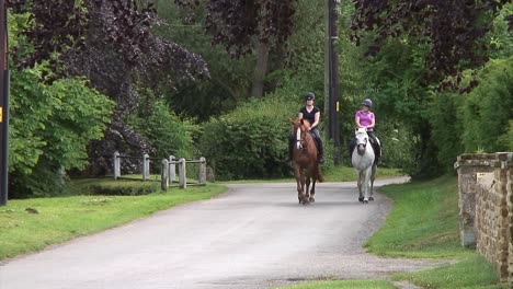 Dos-Mujeres-Jinetes-En-Una-Carretera-Rural-Ejerciendo-Suavemente-Sus-Caballos-En-Una-Hermosa-Tarde-En-La-Campiña-Inglesa