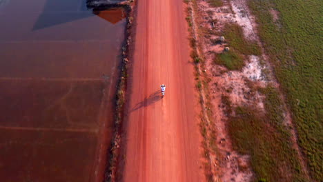 El-Dron-Sigue-A-Un-Motociclista-En-Un-Polvoriento-Camino-De-Tierra-Entre-Granjas-Y-Plantaciones-De-Sal.