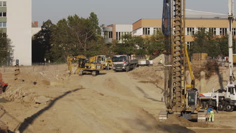 Industrial-drill-in-the-foreground-with-people-around-it-and-two-excavators-in-the-background-scooping-dirt-on-a-construction-site-in-Vienna,-Austria