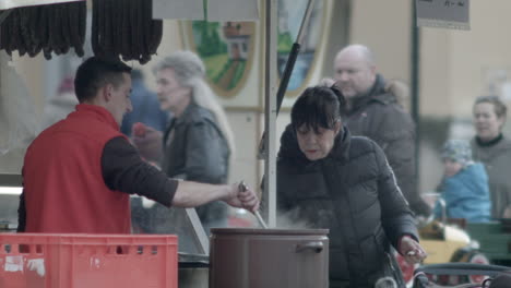 A-woman-enjoys-food-from-a-street-vendor's-large-pot-as-people-walk-behind-at-the-Slavic-Carnival-in-Melnik,-Czech-Republic