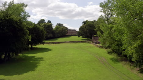 Aerial-Reveal-Shot-of-a-Country-Manor-House-nestled-among-a-Tree-Lined-Avenue-in-England,-UK-on-a-Sunny-Summer’s-Day-with-Blue-Sky---White-Clouds