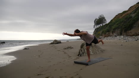 Ein-Yogalehrer-Eines-Jungen-Mannes,-Der-Schwierige-Balancierungsstellungen-Am-Strand-Mit-Meereswellen-In-Zeitlupe-Macht