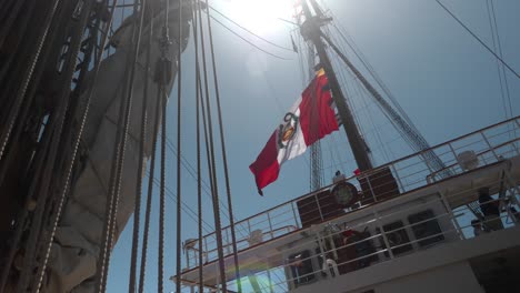 Sun-flair-shines-through-tall-ship-ropes-and-rigging-as-large-colorful-flag-waves-in-breeze