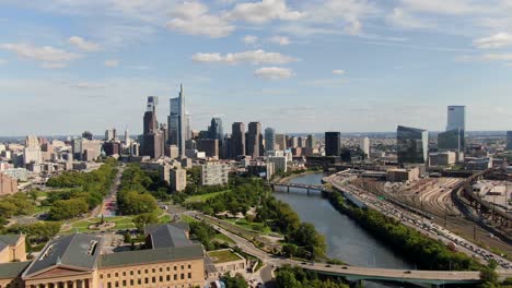 Toma-Aérea-De-Un-Dron-Volador-Hacia-Atrás-Que-Revela-El-Museo-De-Arte-De-Filadelfia-Junto-Al-Río-Schuylkill-Con-El-Hermoso-Horizonte-De-Filadelfia-Contra-El-Cielo-Azul-De-Verano