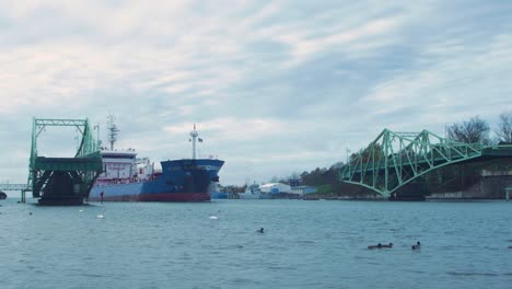 Blaues-Frachtschiff-Fährt-An-Bewölktem-Herbsttag-über-Eine-Offene-Oskara-kalpaka-drehbrücke-In-Liepaja-In-Den-Hafen-Ein,-Wildenten-Im-Vordergrund,-Weitschuss