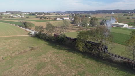 Vista-Lateral-Aérea-De-Una-Antigua-Locomotora-De-Vapor-Restaurada-Que-Retrocede-En-El-Campo-Que-Sopla-Humo-Negro-Y-Vapor