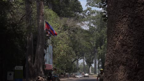 Amplio-Plano-Exterior-De-La-Carretera-Una-Bandera-Camboyana-Voladora-Mientras-El-Hombre-Sale-De-Detrás-Del-árbol-Y-Camina-Hacia-La-Cámara-Con-Casco-De-Seguridad