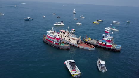 Flotilla-of-boats-surrounding-a-ship-preparing-to-be-sunk,-aerial-POI