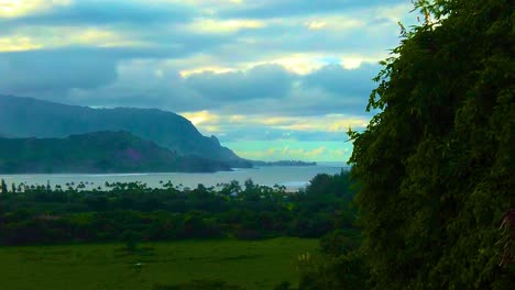 HD-Hawaii-Kauai-slow-motion-pan-right-to-left-of-greenery-in-frame-right-with-a-cove-and-mountainous-coastline-in-the-distance-ending-with-sunlight-through-clouds