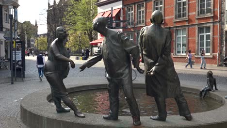 Estatua-De-Bronce-Con-Gente-Y-Piscina-De-Hidromasaje-En-Aquisgrán,-Alemania,-Llamada-Circulación-De-Dinero-Con-La-Catedral-De-Aachener-Dom-En-Segundo-Plano