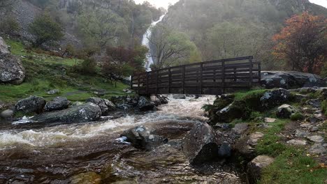 Slow-motion-powerful-rapid-flowing-water-cascades-under-wooden-bridge-from-woodland-valley-waterfall-wilderness