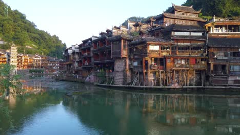 Morning-view-of-the-sightseeing-old-wooden-tourist-boat-sailing-among-the-ancient-stilt-wooden-houses-built-on-the-riverbanks-of-the-Tuo-river,-flowing-through-the-centre-of-Fenghuang-Old-Town