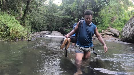 Un-Guía-Local-En-Kokoda,-Papúa-Nueva-Guinea,-Está-Cruzando-Un-Río-Que-Le-Llega-Hasta-Las-Rodillas-Con-Su-Mochila-Y-Su-Machete-Firmes-Y-Con-Cuidado