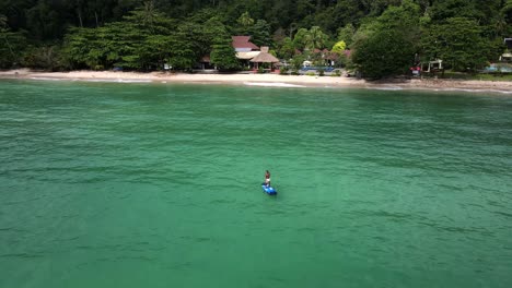 Aerial-drone-bird's-eye,-backwards-dolly-shot-of-Asian-man-exercising-on-a-sup-paddle-board-in-turquoise-tropical-clear-waters,-with-beach,-jungle-,-resorts-and-coastline-in-Thailand