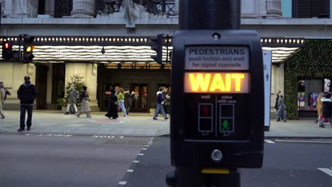 A-slow-motion-left-to-right-truck-camera-technique-past-a-pedestrian-crossing-signal-to-reveal-a-people-walking-past-a-social-distancing-sign-on-front-of-Selfridges-store-entrance-on-Oxford-Street