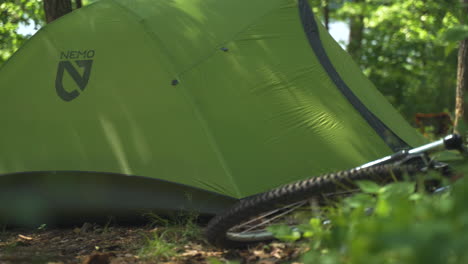 Una-Carpa-Nemo-Verde-Brillante-Se-Encuentra-En-Medio-De-Un-Bosque-Arbolado-Soleado-Junto-A-Una-Bicicleta-De-Montaña-Durante-Un-Viaje-De-Campamento-De-Verano