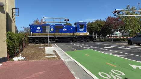 Blue-train-caboose-passes-over-city-street-behind-lowered-crossing-gate