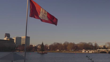 The-flag-of-Hamburg-flapping-in-front-of-the-Christmas-tree-floating-at-Binnenalster-in-Hamburg,-Germany,-in-Dec-2019
