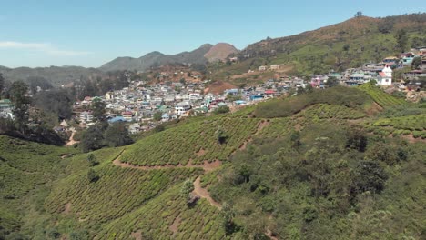 Rural-village-settled-on-the-slope-of-a-hill-surrounded-by-Tea-Gardens-in-Munnar,-India---Aerial-wide-Establishing-shot