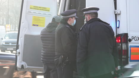 Police-officers-in-face-masks-search-a-detained-man-retrained-in-handcuffs-next-to-a-police-van,-who-is-suspected-of-breaching-Coronavirus-legislation