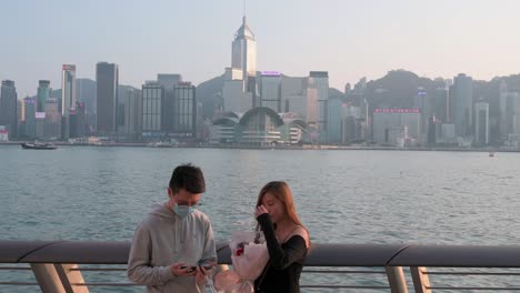 Una-Pareja-Toma-Una-Foto-Mientras-Sostiene-Una-Boutique-De-Rosas-A-Lo-Largo-Del-Paseo-Marítimo-Del-Puerto-Victoria-Y-El-Horizonte-De-La-Isla-De-Hong-Kong-En-El-Fondo-Mientras-Se-Pone-El-Sol