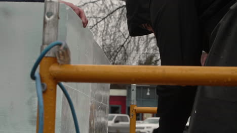 Tilt-down-on-ice-sculptor-cutting-through-ice-block-with-reciprocating-saw-in-falling-snow,-Slow-Motion