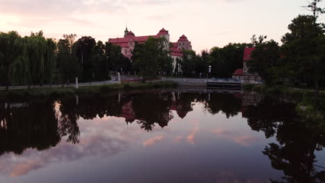 Revealing-14th-century-palace-in-Niemodlin-from-looking-down-at-pond-with-still-water-reflecting-the-sky,-clouds,-trees,-and-the-palace-itself