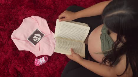 Una-Mujer-Embarazada-Disfruta-Sentada-Leyendo-Un-Libro,-Frente-A-Ella-Hay-Ropa-De-Bebé-Rosa-Y-Una-Fotografía-De-Ultrasonido