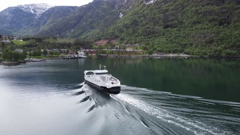 Battery-powered-ferry-Kinsarvik-from-Boreal-company-heading-for-port---Aerial-passing-around-stern-of-ship-sailing-full-ahead-with-Kinsarvik-town-and-mountain-landscape-in-background---Norway