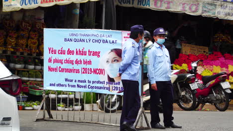 Statische-Ansicht-Von-Polizisten,-Die-Vor-Einem-Plakat-Sprechen,-Das-Sich-Auf-Covid-Regeln-In-Der-Straße-In-Der-Nähe-Des-Marktes-Da-Lat-Bezieht