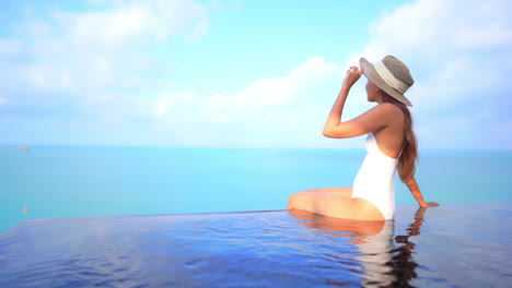Fitte-Asiatische-Frau-Im-Weißen-Badeanzug,-Die-An-Einem-Sonnigen-Tag-In-Zeitlupe-Am-Rand-Des-Infinity-Pools-Auf-Dem-Dach-Sitzt,-Auf-Einem-Atemberaubenden-Türkisfarbenen-Meerblick-Hintergrund-Auf-Den-Bahamas