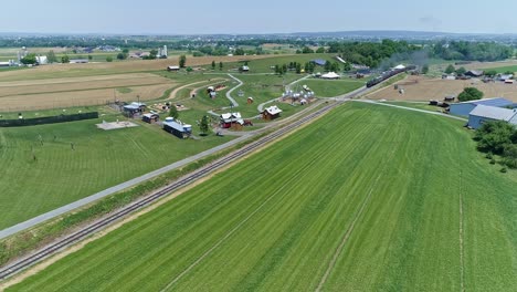 An-Aerial-View-of-a-Steam-Engine-Puffing-Smoke-and-Steam-with-Passenger-Coaches-Approaching-Passing-a-Corn-Maze-Park-and-Farmland-Countryside-on-a-Beautiful-Cloudless-Spring-Day