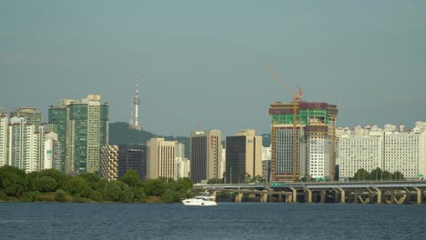 White-Yacht-Boat-Floating-on-Hangang-River-with-Seoul-Cityscape---BBS-Bulgyobangsongguk-building,-Hanwha-Obelisk-apartment,-Mapodaegyo-Mapo-bridge,-Seoul-Namsan-Tower,-Bamseom-Island-panoramic-view