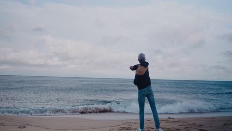 Young-Tall-Guy-With-Jacket-Throwing-Pebbles-and-Rocks-Into-The-Ocean---Static-Wide-Shot