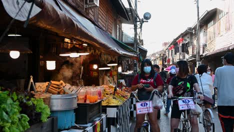 Two-ladies-on-their-bicycles-stopping-to-buy-some-street-Thai-food-along-the-Walking-Street,-Chiang-Khan-in-Loei,-Thailand