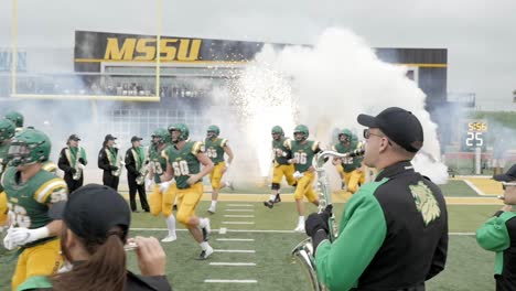 Slow-motion-college-football-players-break-through-paper-pregame-and-run-onto-the-field-as-champions-while-marching-band-and-cheerleaders-cheer-Stock-Video-Footage