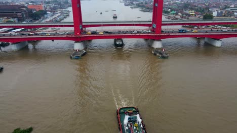 Industrial-boats-sailing-under-Ampera-bridge-in-Palembang-city,-aerial-tilting-up-view