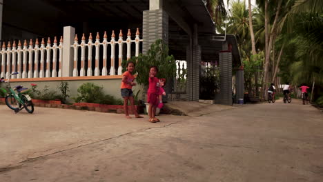 Two-Asian-Little-Girls-in-Rural-Village-Smiling-at-Camera,-One-Child-Holding-Doll-Waving