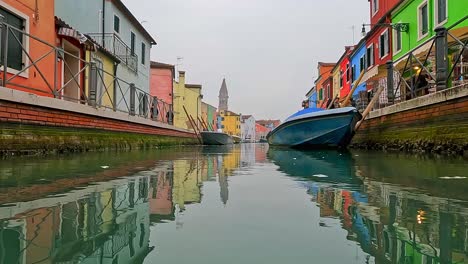 Panorámica-De-La-Superficie-Del-Agua-Vista-Pov-De-Casas-Coloridas-De-Burano-Y-Canal-Con-Barcos-Atracados,-Italia