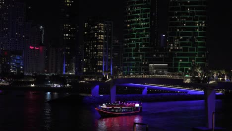 Dubai-Marina-Bei-Nacht,-Beleuchtetes-Boot-Mit-Neonlichtern,-Die-Im-Kanal-Unter-Wolkenkratzern-Segeln